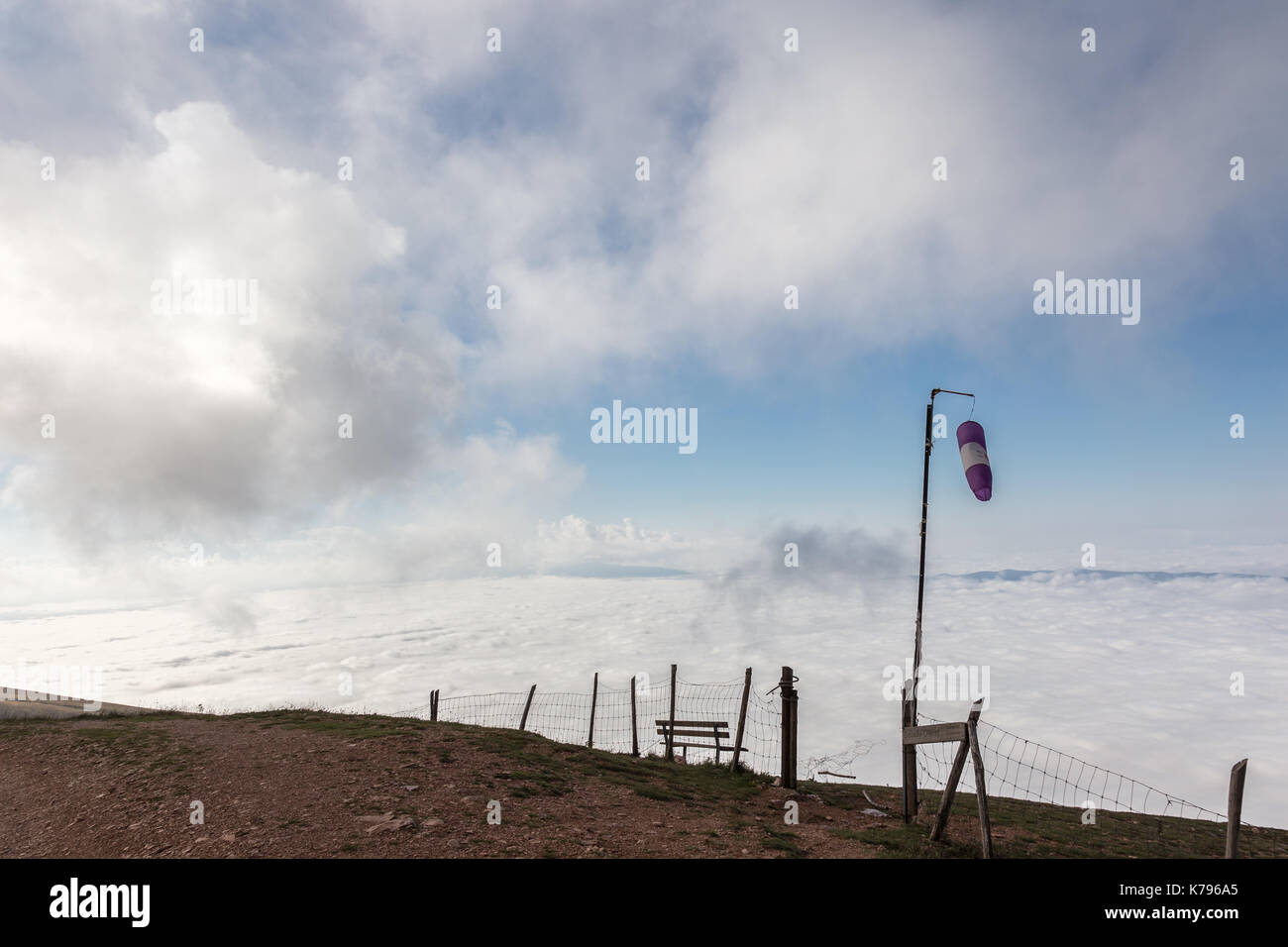 A mountain top over a valley filled by fog, with very low clouds, fence and a windswept windsock Stock Photo