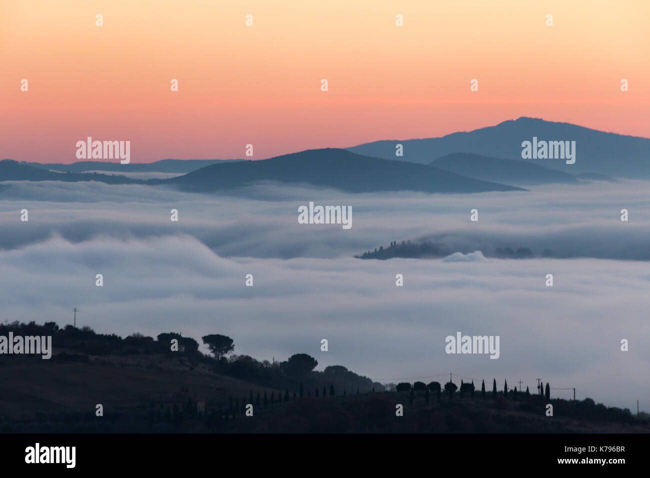 A valley filled by fog at sunset, with some hills and trees in the foreground and other hills and mountains in the foreground Stock Photo