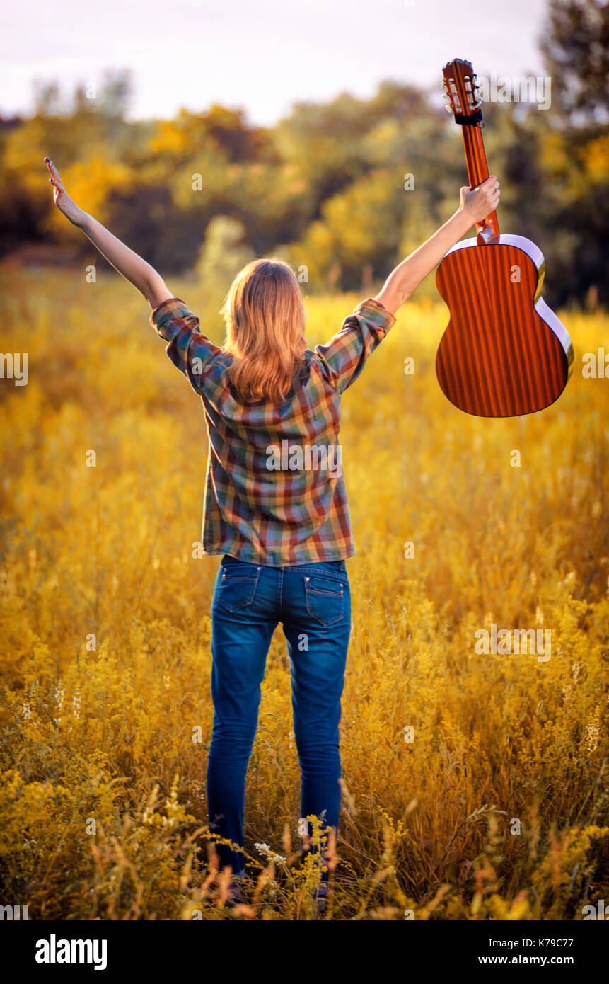 Image from the back of a happy young woman on the plaid shirt and blue jeans.  Hipster stand up hands  to sky with a acoustic guitar on a  yellow autu Stock Photo