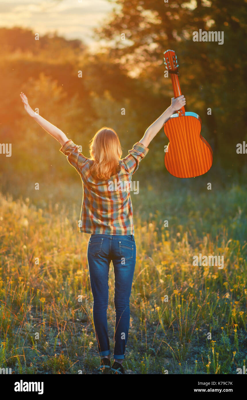 Image from the back of a happy young woman on the plaid shirt and blue jeans.  Girl stand up hands  to sky with a acoustic guitar on a  yellow autumn  Stock Photo