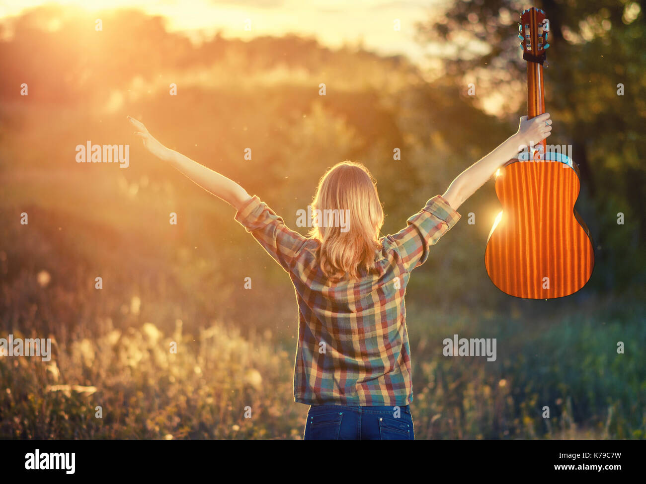 young girl in a plaid shirt at sunset with an acoustic guitar. A woman rejoicing in the autumn weather raised her hands up. Backlight Stock Photo