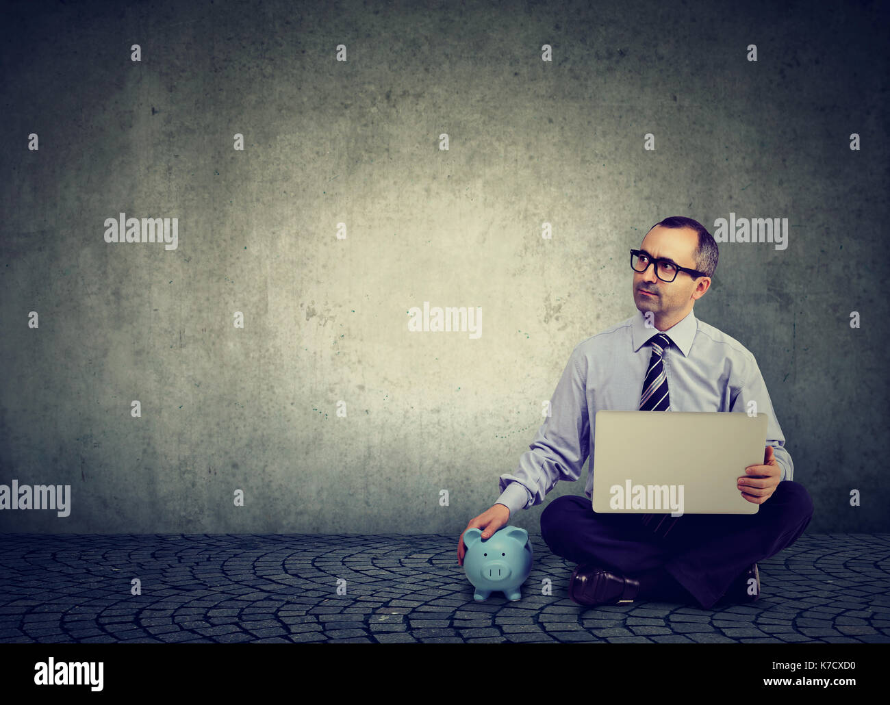 Thoughtful business man with laptop and piggy bank sitting on a floor Stock Photo