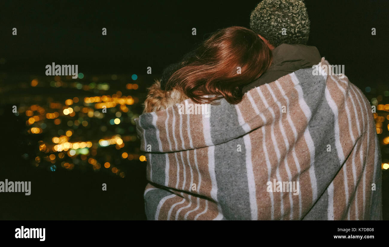 Rear view of young couple wrapped in a blanket against cityscape during night Stock Photo