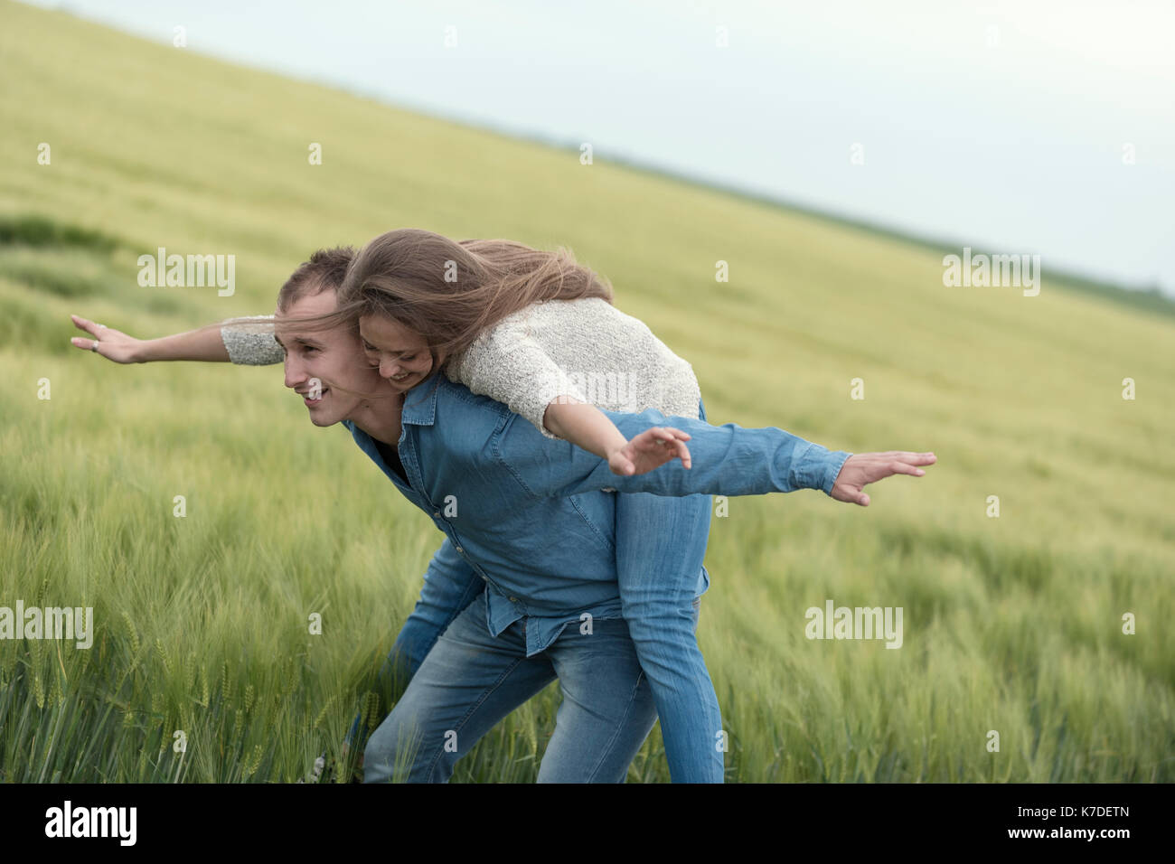 Cheerful couple with arms outstretched enjoying piggyback ride on grass field against sky Stock Photo