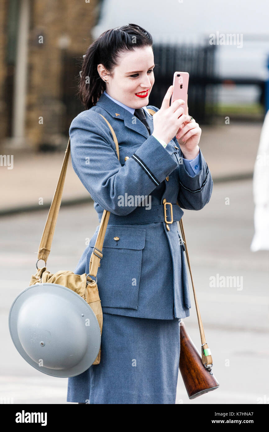 England, Chatham Dockyard. Event, Salute to the forties. Women in RAF uniform with rifle slang over shoulder and tin helmet hanging from other shoulder. Standing, smiling and using iPhone to film event. Stock Photo