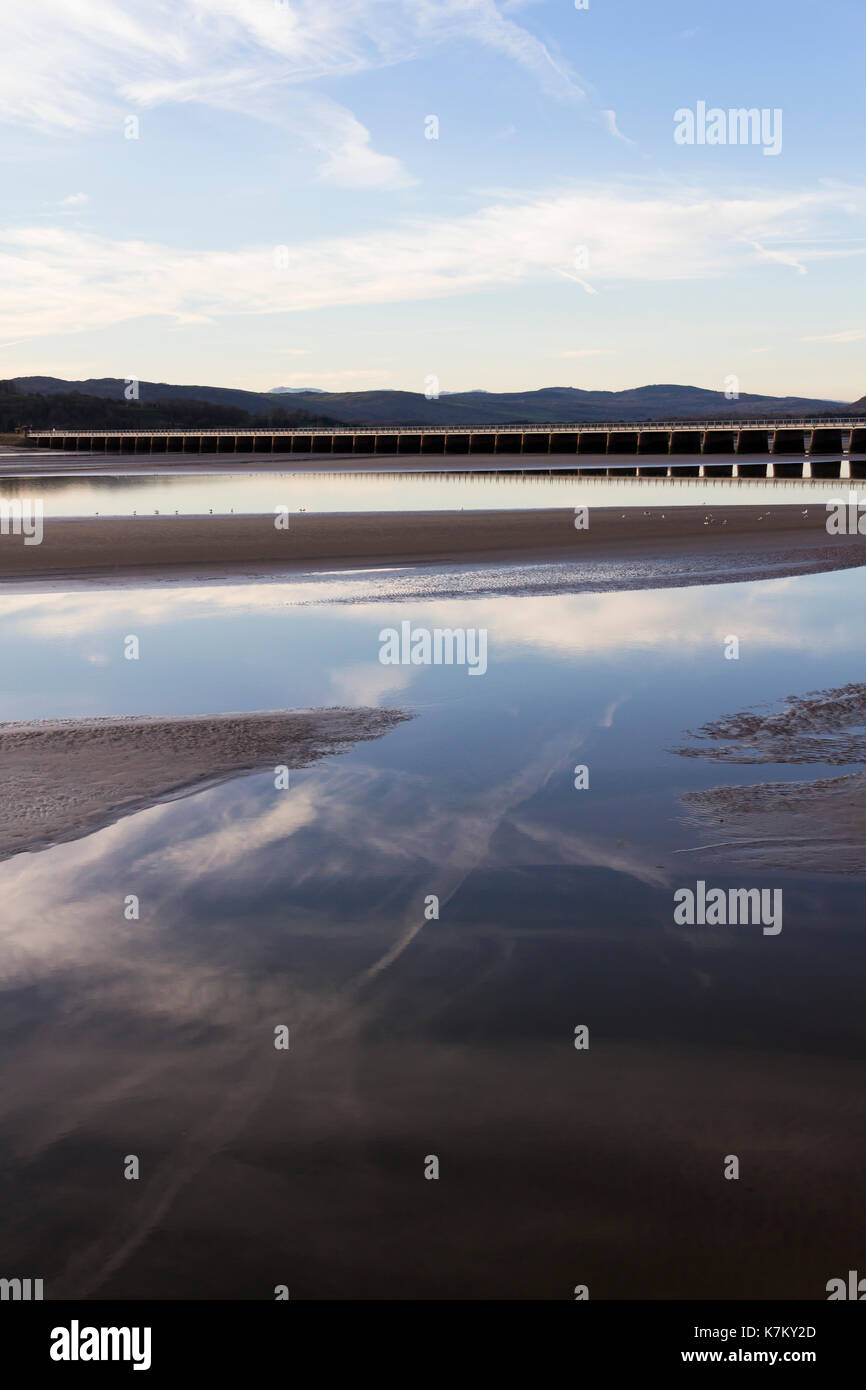 Reflected vapour trails in the calm surface of the River Kent estuary at low tide, with the Arnside railway viaduct beyond. Stock Photo