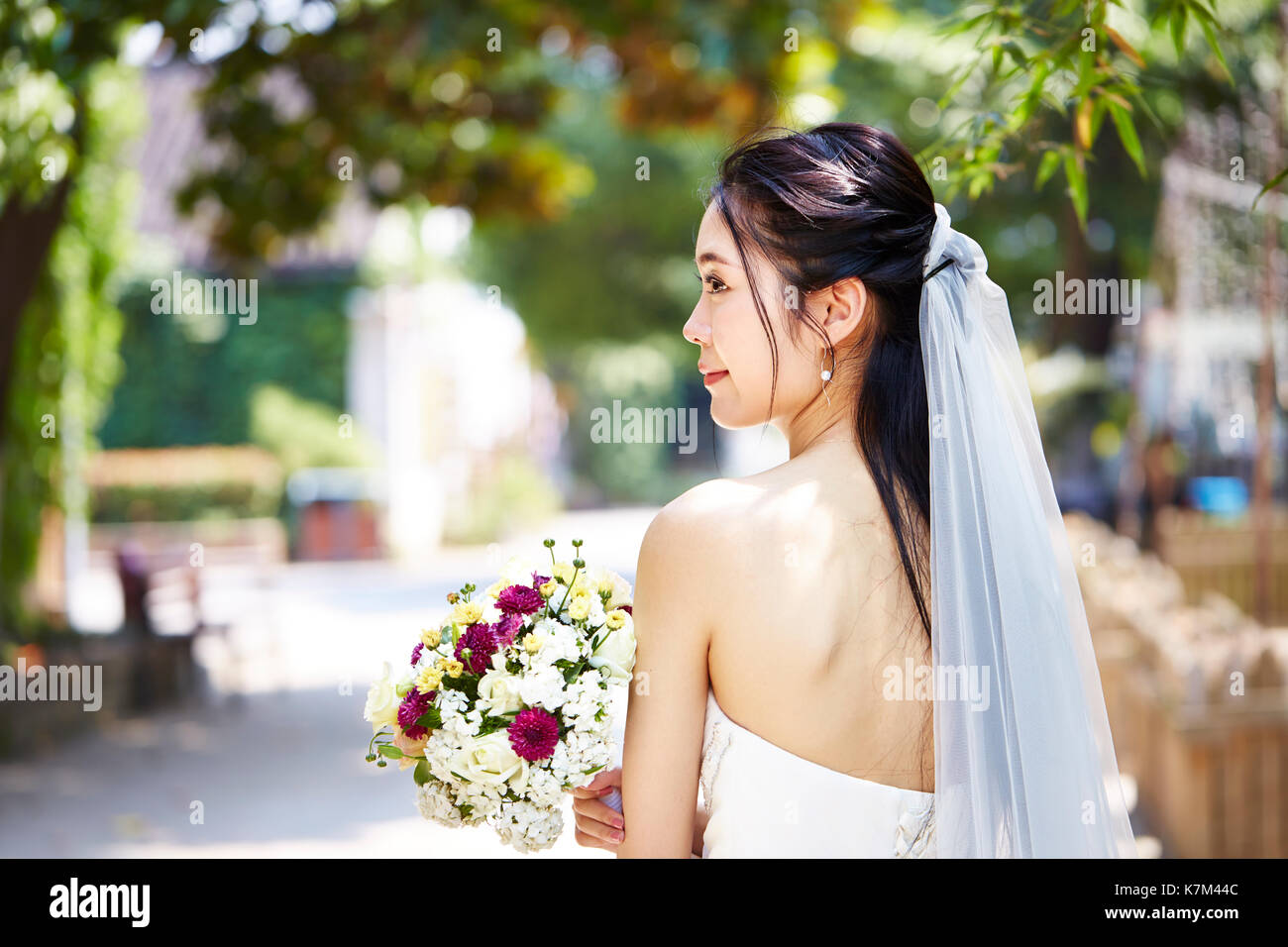 beautiful and happy young asian bride wearing bridal veil holding a bunch of flower, rear view. Stock Photo