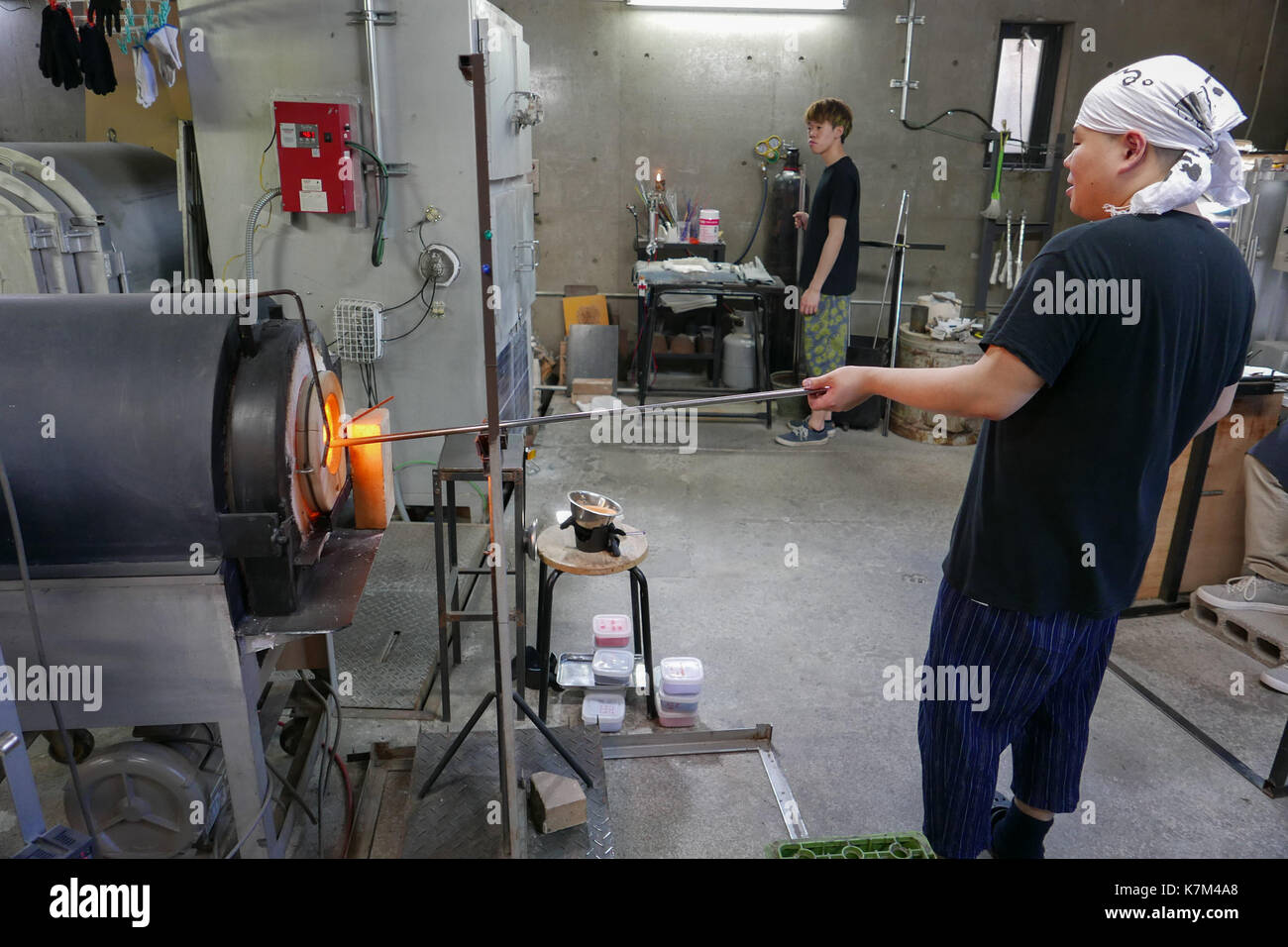 White bandana wearing Japanese glass work craftsman holding long metal rod in glass furnace with workshop in the background.  Yellow flame and heat. Stock Photo