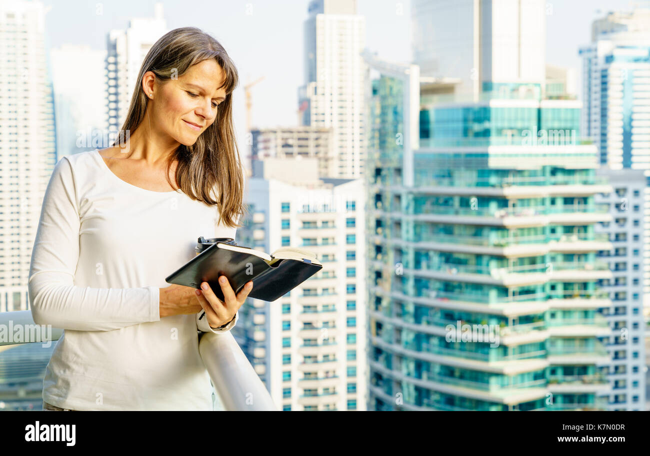 Mature woman is reading a book on her balcony in a highrise building Stock Photo