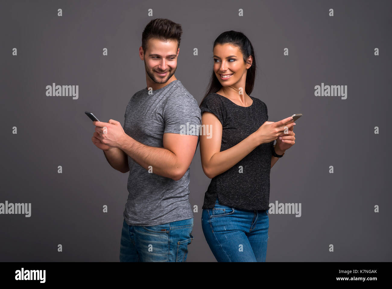 A beautiful young couple standing back to back while holding and using their smartphones Stock Photo