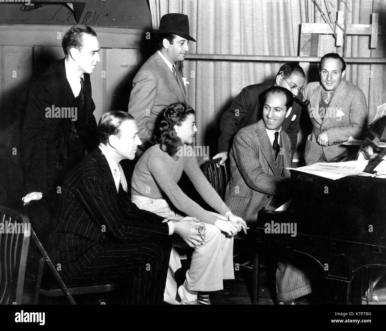 On the movie set of 'Shall We Dance,' George Gershwin is photographed at the piano with (l-r) Hermes Pan, dance director; Fred Astaire; Mark Sandrich, director of the film; Ginger Rogers; Mr. Gershwin; his brother Ira Gershwin, and Nathaniel Shilkret, musical director, Hollywood, CA, 1936. Stock Photo