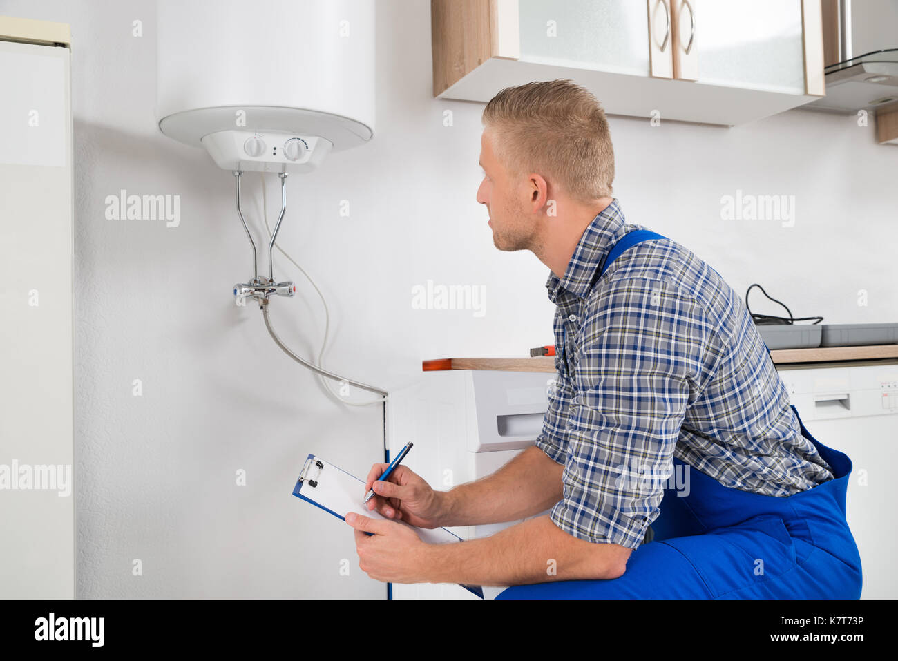Male Plumber Writing On Clipboard In Front Of Electric Boiler In Kitchen Room Stock Photo