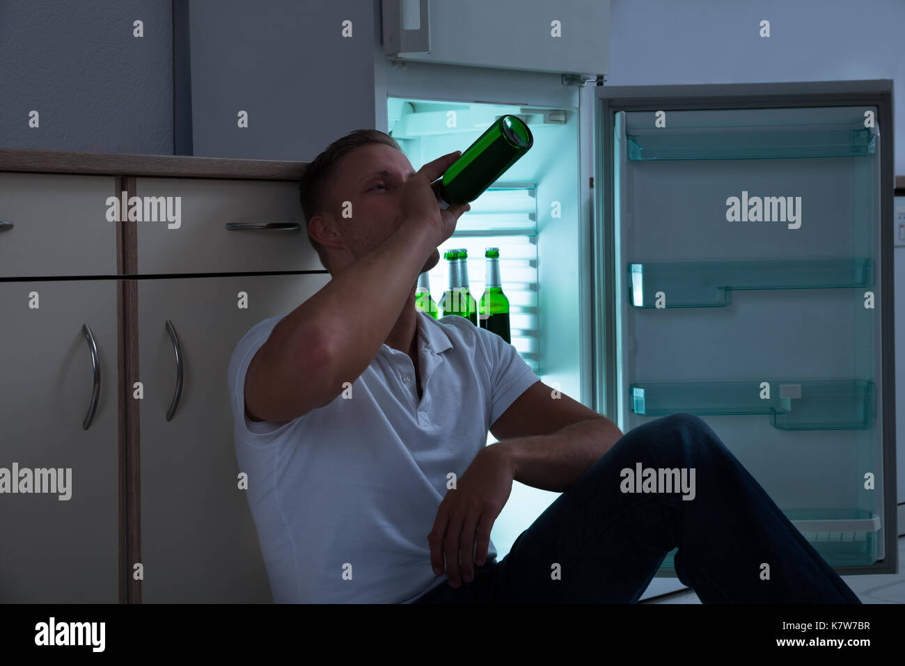Young Man Sitting Near The Cabinet And Drinking Beer In Kitchen Stock Photo