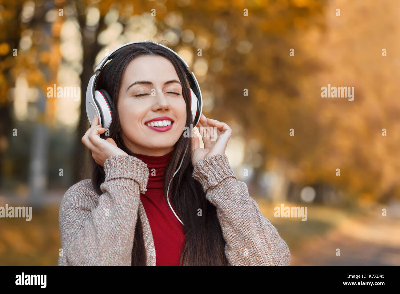 young smiling woman in headphones with closed eyes outdoors on autumn day. Girl listening music in park. Girl enjoys music. Autumn portrait Stock Photo