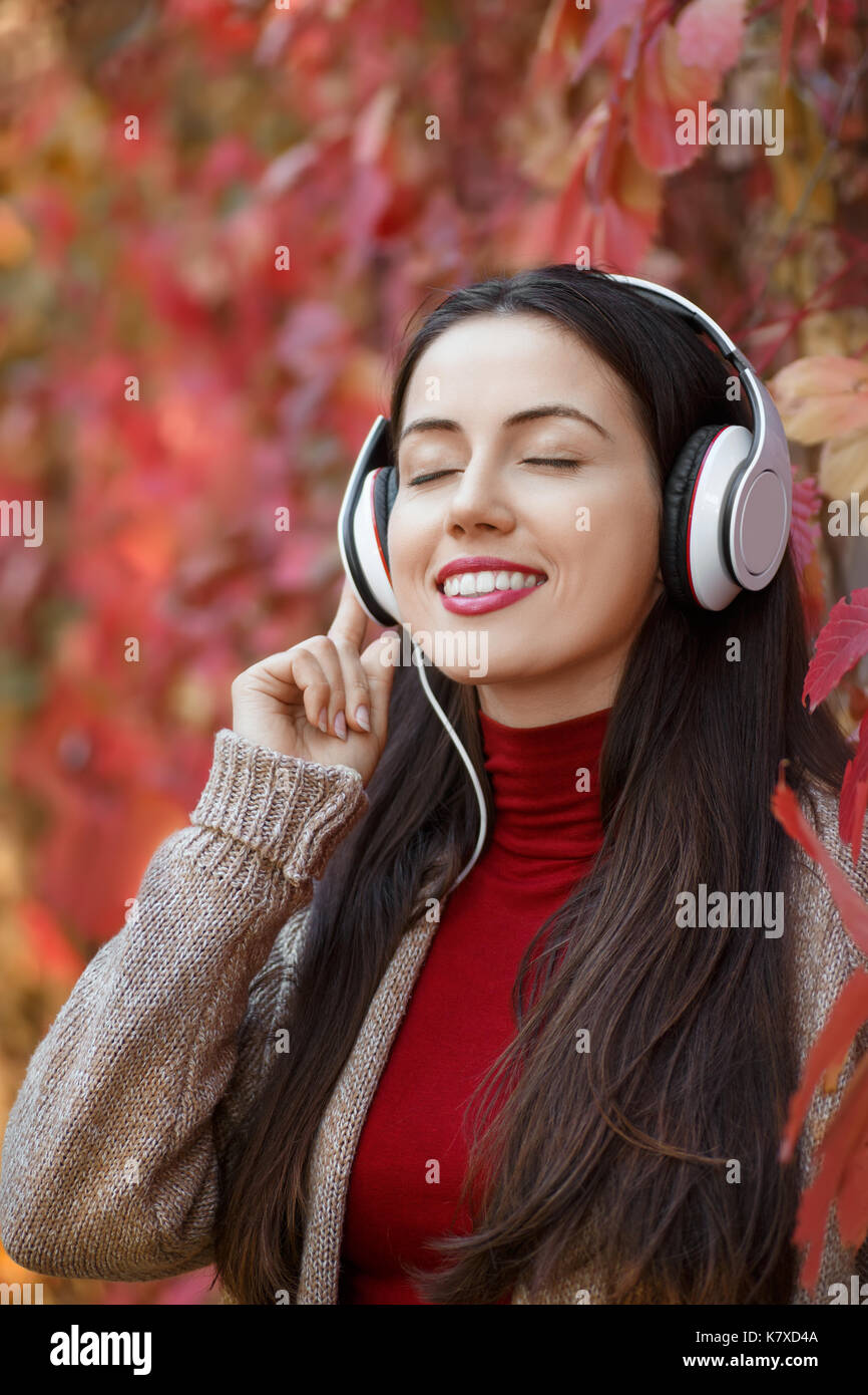 young smiling woman in headphones with closed eyes outdoors on autumn day. Girl listening music in park. Girl enjoys music. Autumn portrait Stock Photo