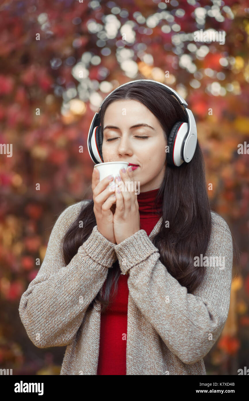 young woman drinking hot takeaway coffee and listening music in autumn park. Girl resting in autumn outdoors Stock Photo