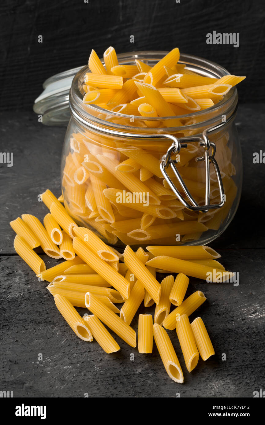 Raw, uncooked, dry penne pasta noodles in glass jar on black table