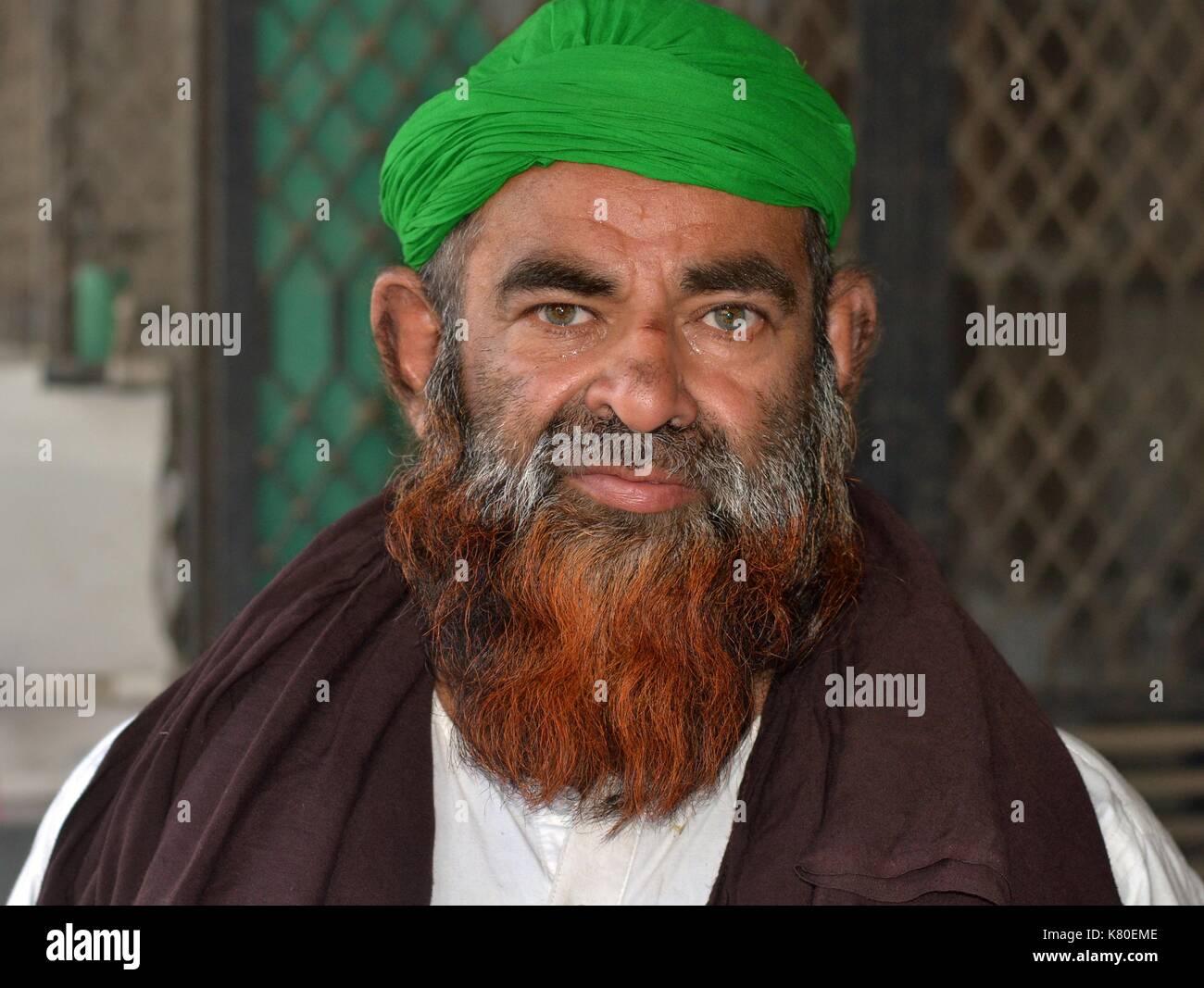 Elderly big Indian Muslim man with henna-dyed Muslim beard wears a green turban and a brown prayer shawl and poses for the camera. Stock Photo