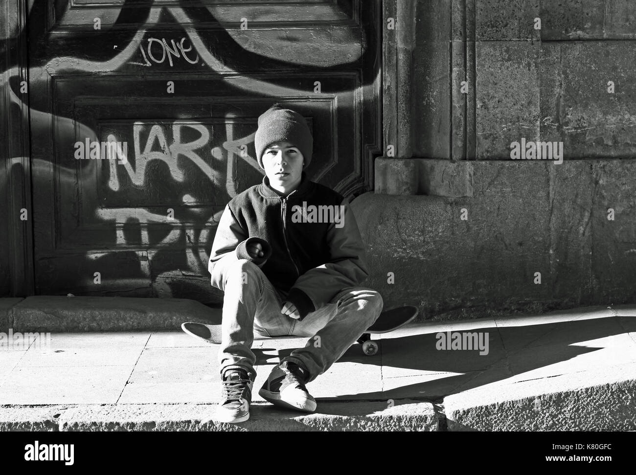 Boy sitting on skateboard in street, in front of graffiti on door of old building in el Raval,  Barcelona Stock Photo