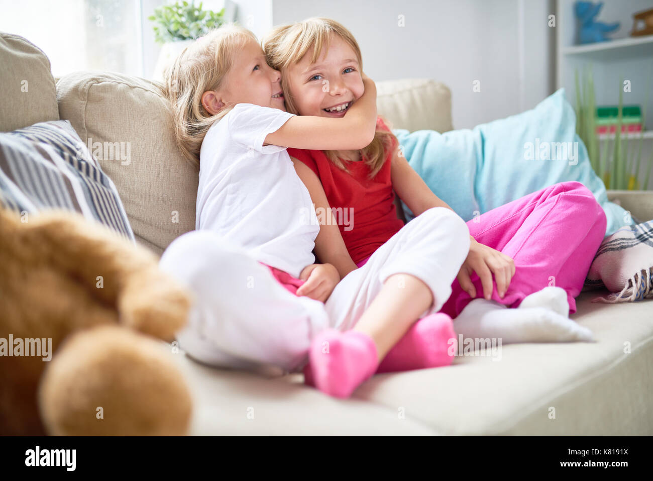 Cute blond-haired girl sharing secret with her smiling elder sister while spending weekend together at cozy living room Stock Photo
