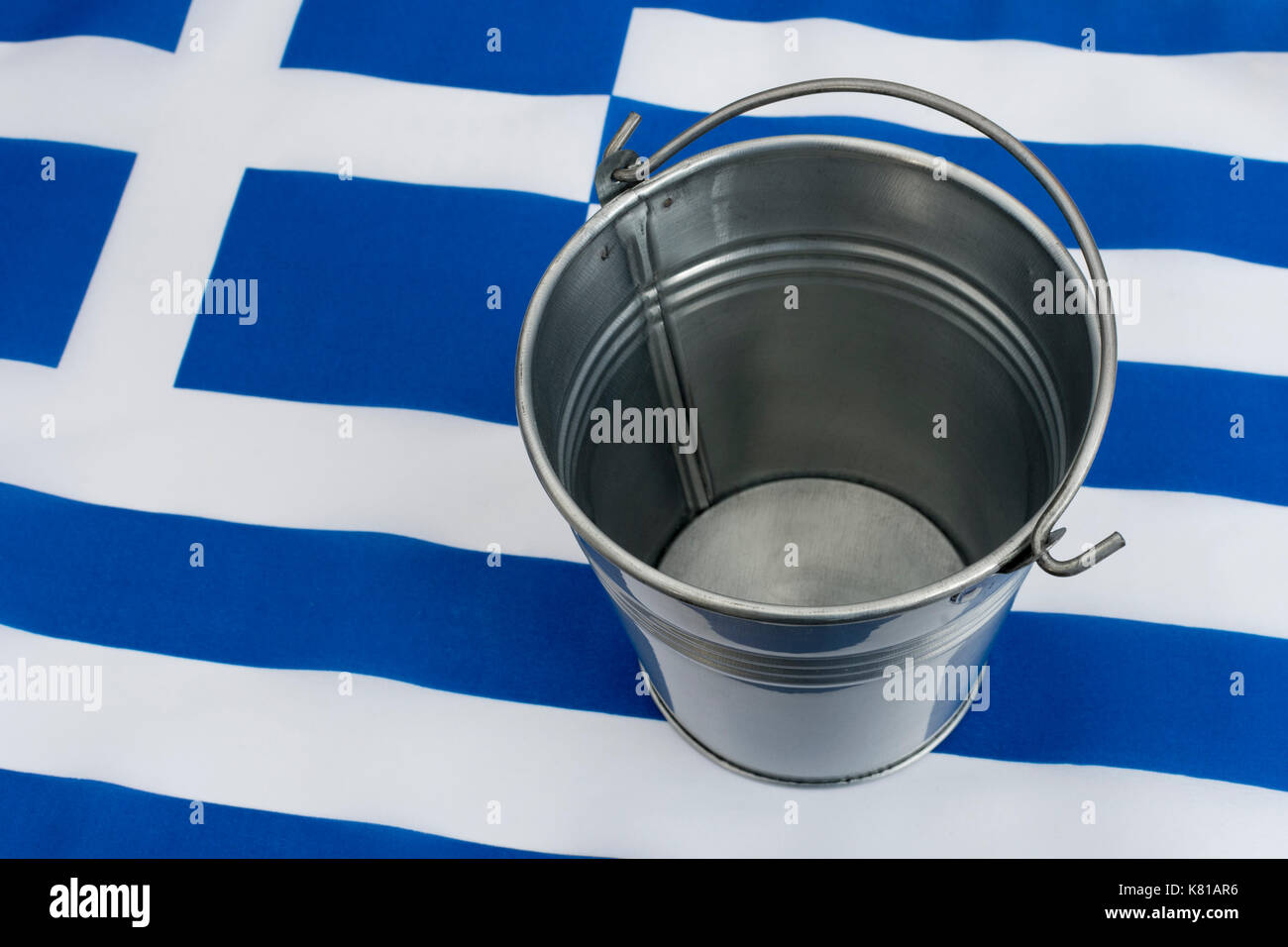Greek flag with an empty metal bucket. For Greek financial crisis & bailout, Greek debt relief, Greece economic slump, Greece recession. Stock Photo