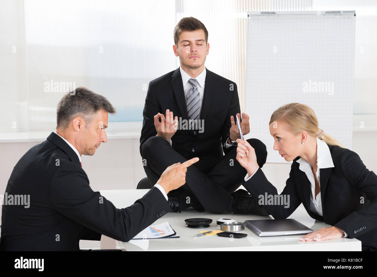 Two Businesspeople Quarreling In Front Of Businessman Meditating At Desk Stock Photo