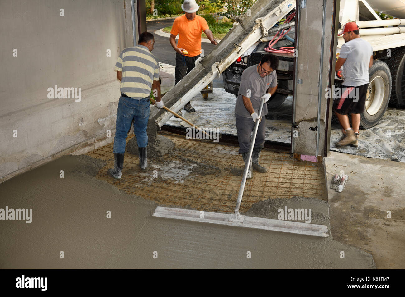 Work crew pouring cement from a cement truck chute over wire mesh on a residential garage floor and spreading with large float trowel and rake Stock Photo