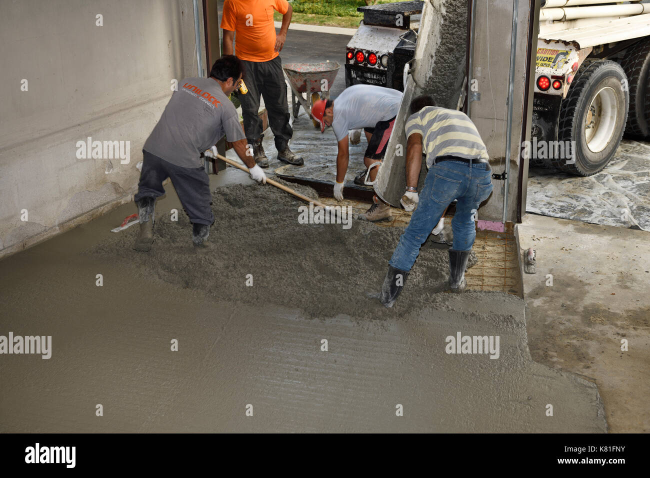 Work crew pouring cement from a cement truck chute over wire mesh on a residential garage floor and spreading with shovel and rake Stock Photo