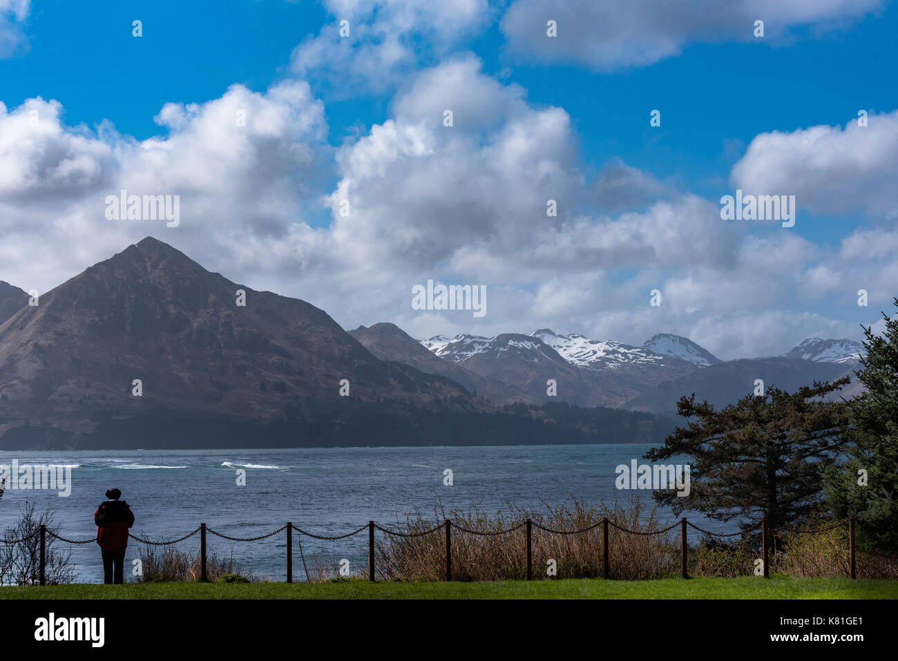 An anonymous man stands by a chain link fence looking at the mountains beyond the sea Stock Photo