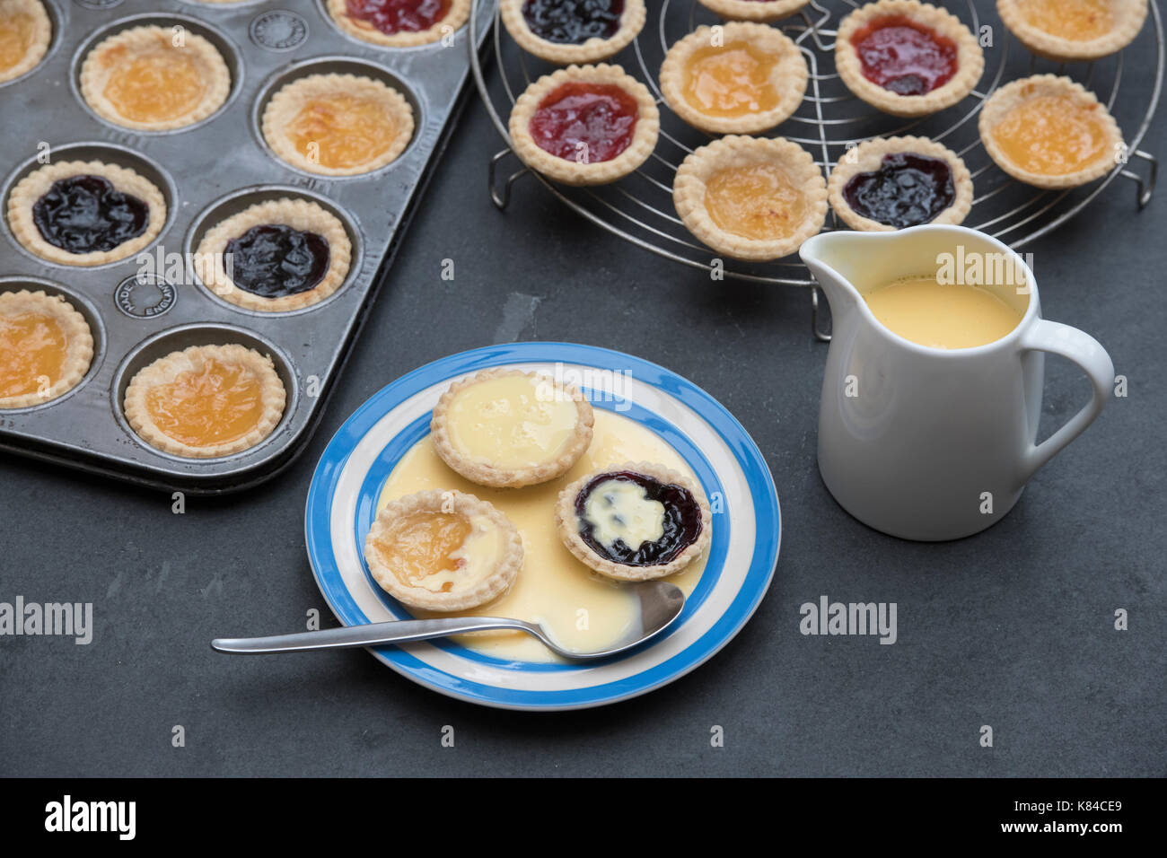 Cooked homemade jam tarts on a plate with custard next to a baking tray and a vintage circular wire rack Stock Photo