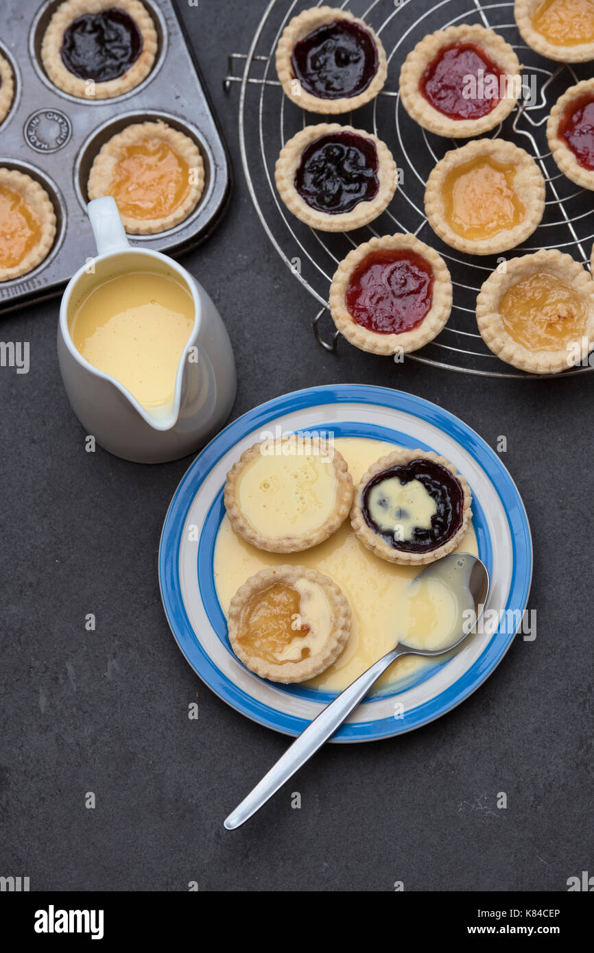 Cooked homemade jam tarts on a plate with custard next to a baking tray and a vintage circular wire rack Stock Photo