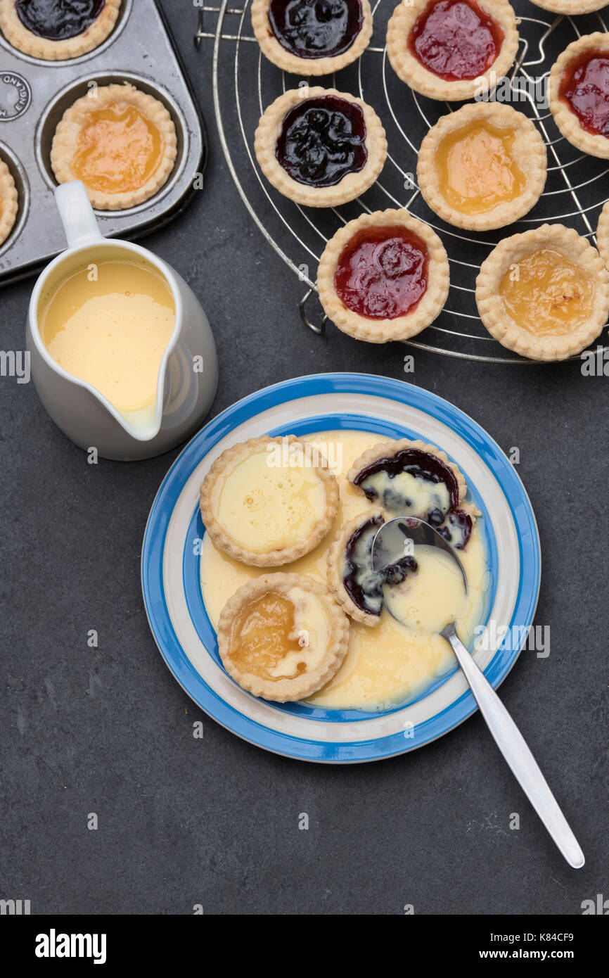 Cooked homemade jam tarts on a plate with custard next to a baking tray and a vintage circular wire rack Stock Photo