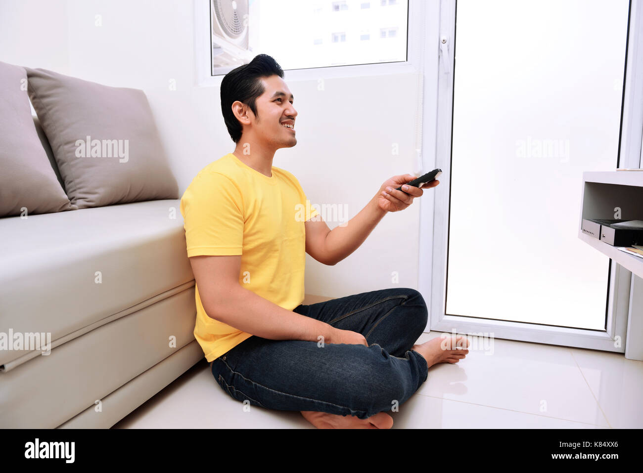 Young asian man holding remote control watching tv while sitting on the floor Stock Photo