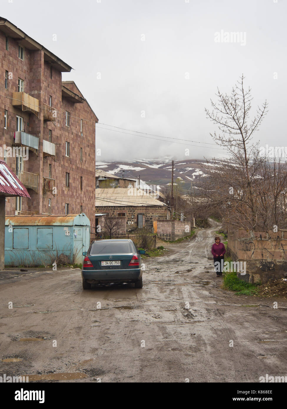 Aparan, a small town in Armenia along the M3 highway, dirt road with rainy potholes, apartment block and view towards the slopes of mount Aragats Stock Photo