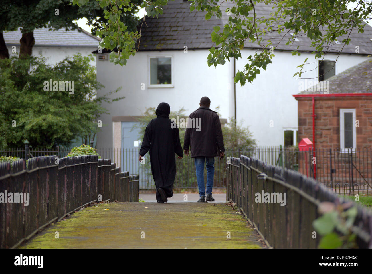 refugee immigrant moslem Muslim couple wearing burka walking on path fenced in, suburban setting Stock Photo