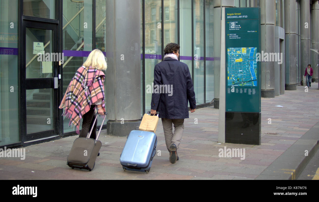 city map man woman couple tourists leaving hotel with trolley cases luggage street Stock Photo