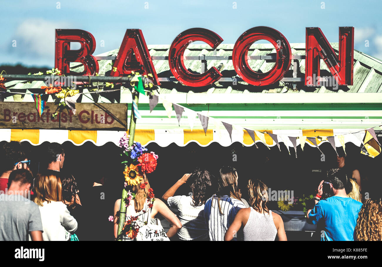 Revellers at a ood stall at a summer music festival red neon sign advertising bacon. Stock Photo