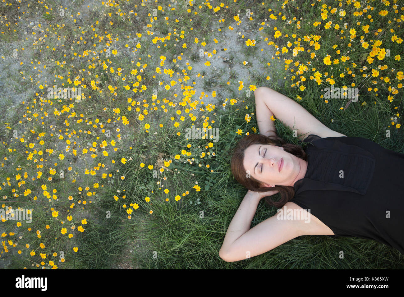 A woman in a black vest top and jeans lying on the grass with her hands behind her head. Stock Photo