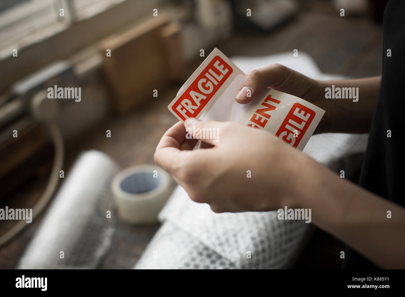 Hands holding a red Fragile sticker to stick it on a brown paper package on a work bench. Stock Photo