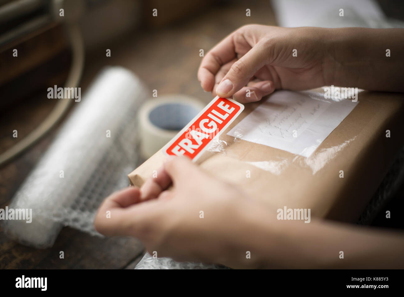 Hands holding a red Fragile sticker to stick it on a brown paper package on a work bench. Stock Photo