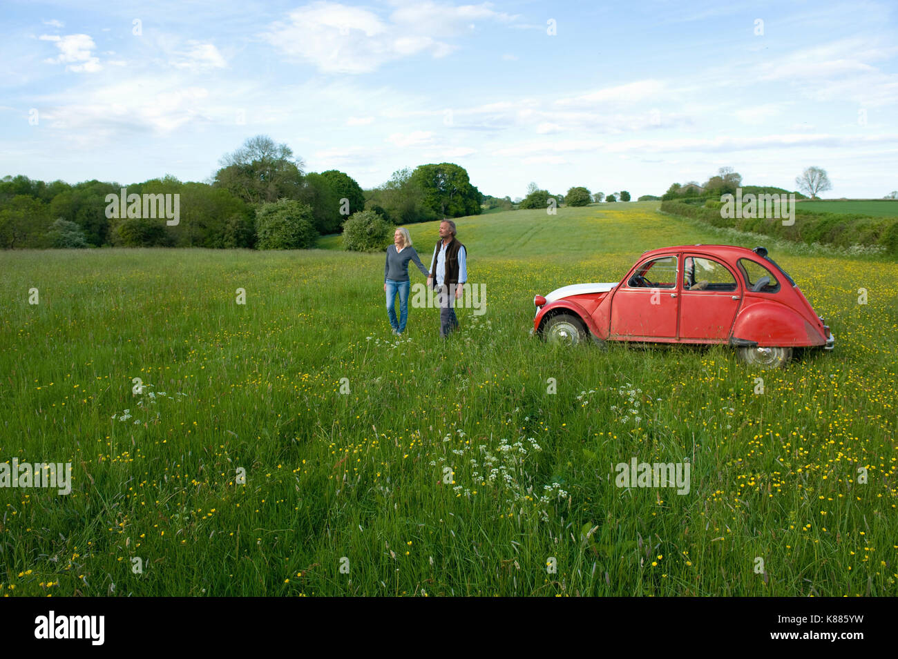 Man and woman walking hand in hand across a meadow, vintage red car parked close by. Stock Photo