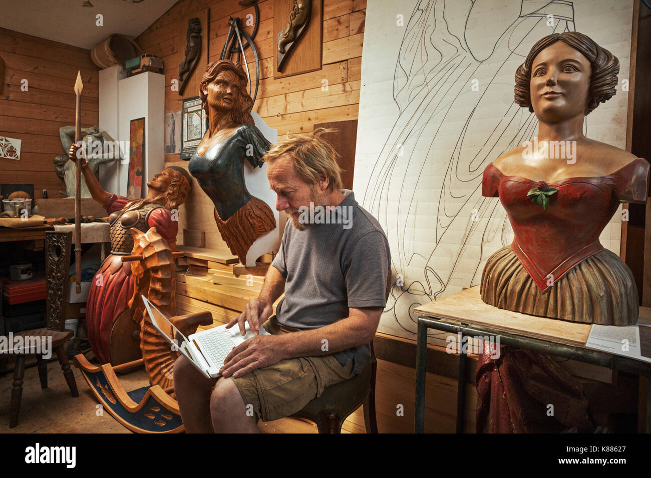 A woodworker, skilled craftsman sitting in a workshop using a laptop on his knees, surrounded by carved wooden female ship's figureheads. Stock Photo
