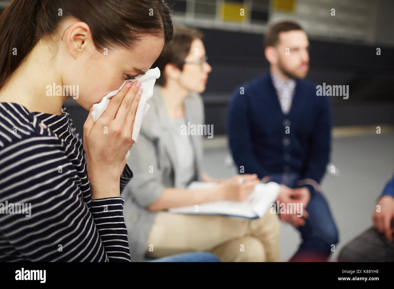 Crying woman wiping her tears with handkerchief with groupmates communicating on background Stock Photo