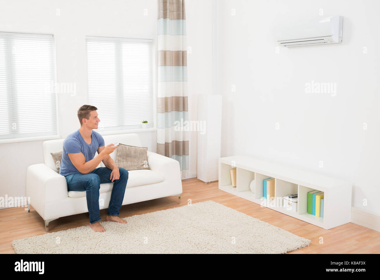Young Man On Sofa Operating Air Conditioner With Remote Control Stock Photo