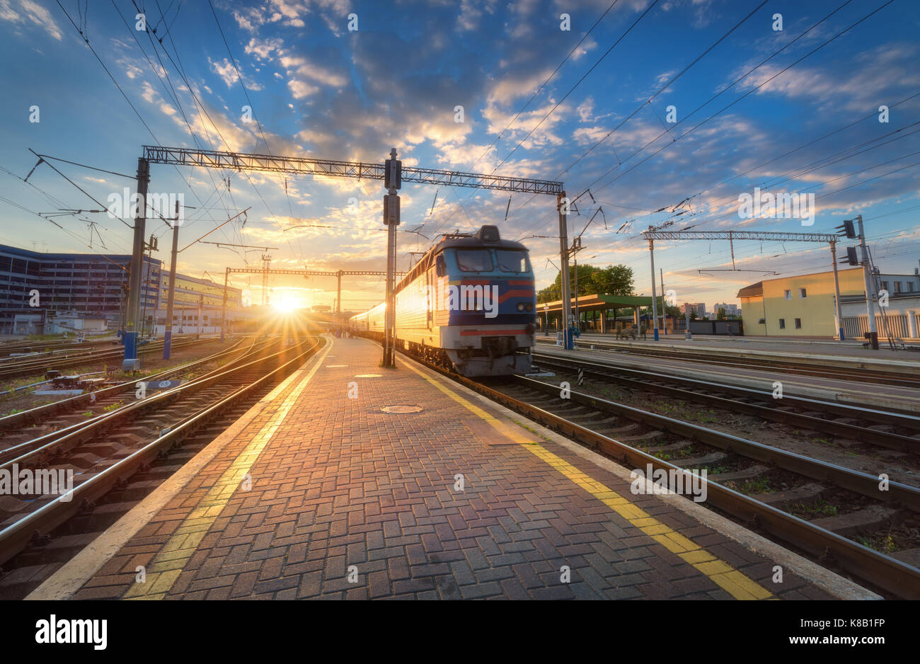 High speed passenger train on railroad track at sunset in Europe. Railway station with modern commuter train departure, blue sky with clouds and sun.  Stock Photo