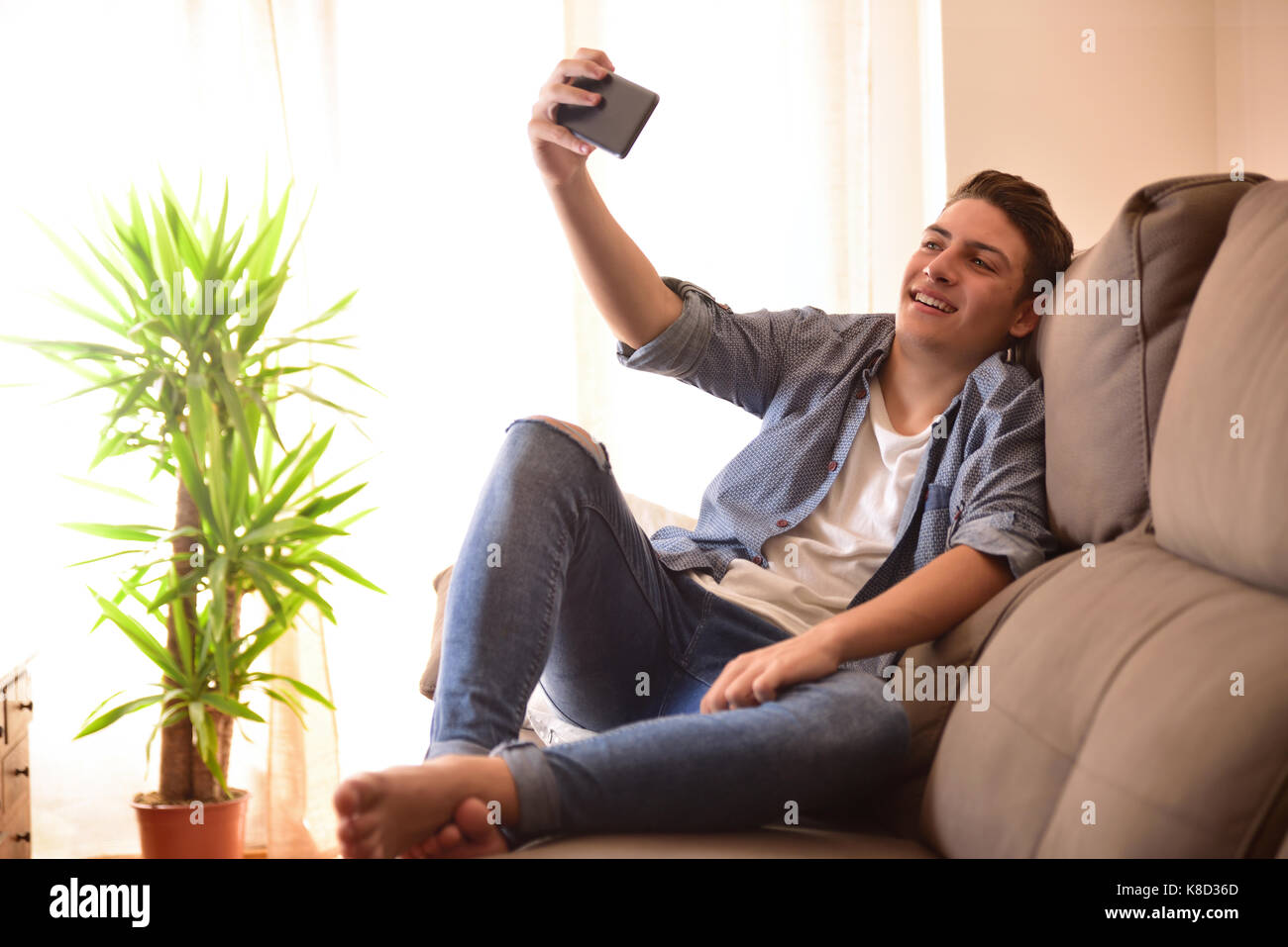 Smiling modern teenager taking a selfie sitting on a sofa in the living room of his house Stock Photo