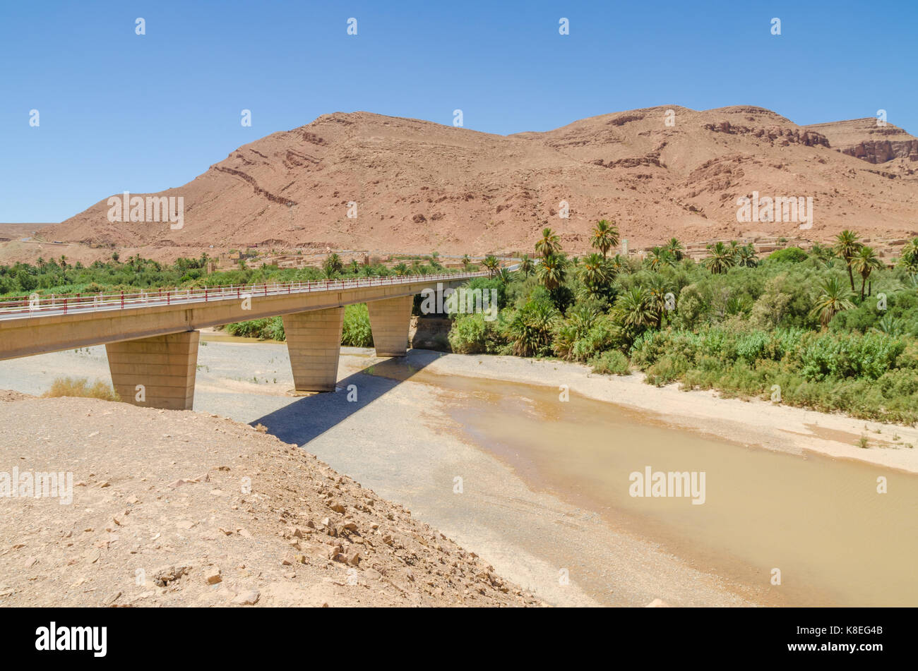 Bridge spanning over dry river bed with some water, mountains and palms in Morocco, North Africa. Stock Photo
