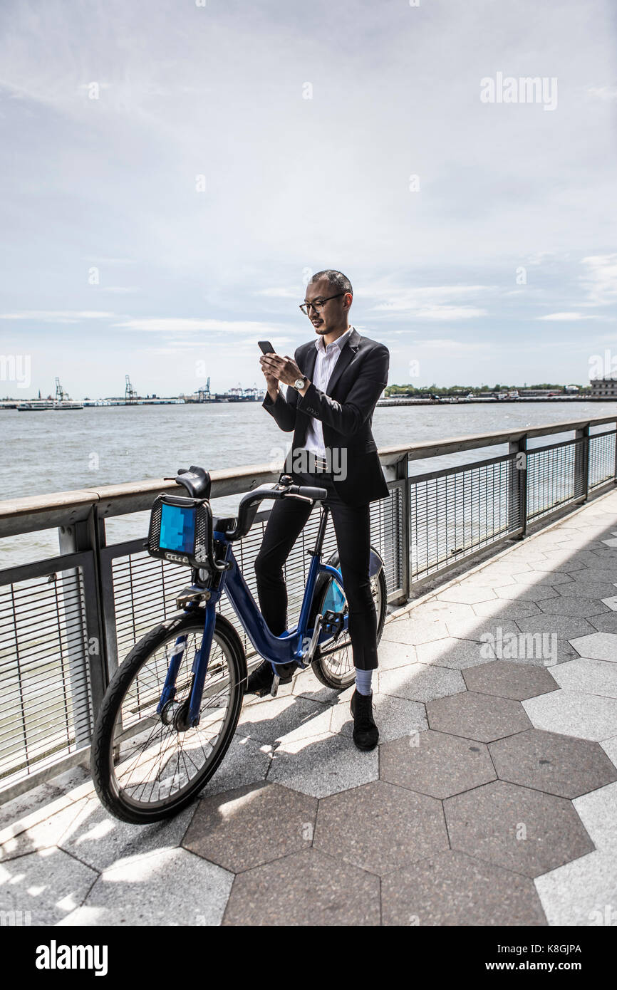 Mid adult businessman on bicycle looking at smartphone along city river waterfront Stock Photo