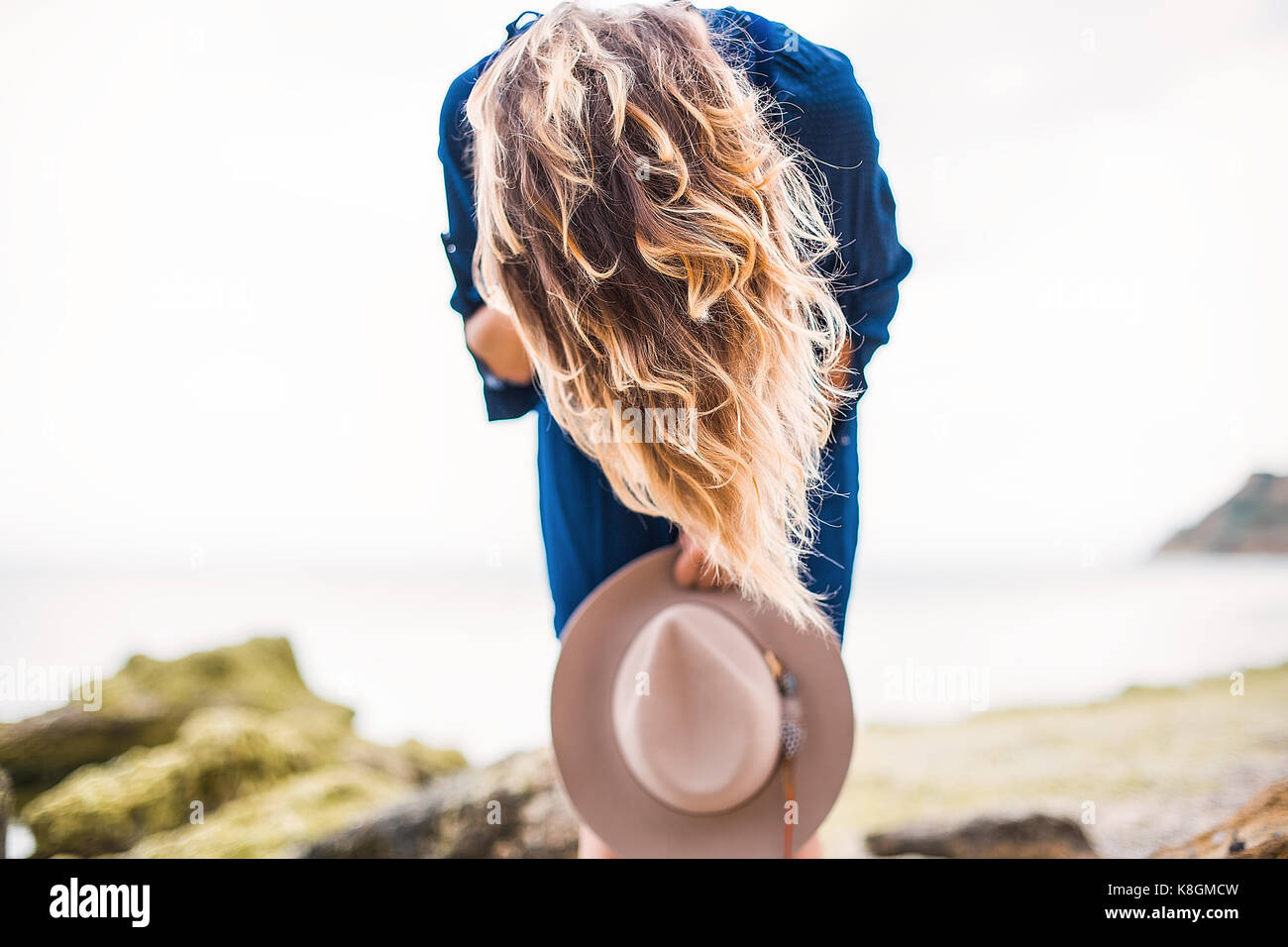Mid adult woman in coastal setting, holding hat, bending over, hair covering face Stock Photo