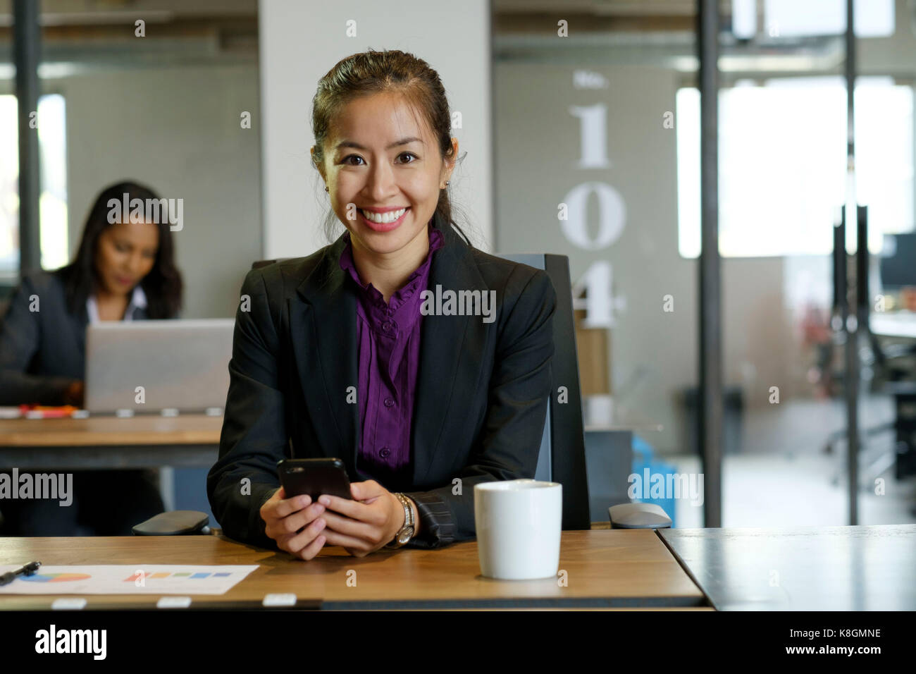Portrait of businesswomen sitting at desk, holding smartphone, smiling Stock Photo